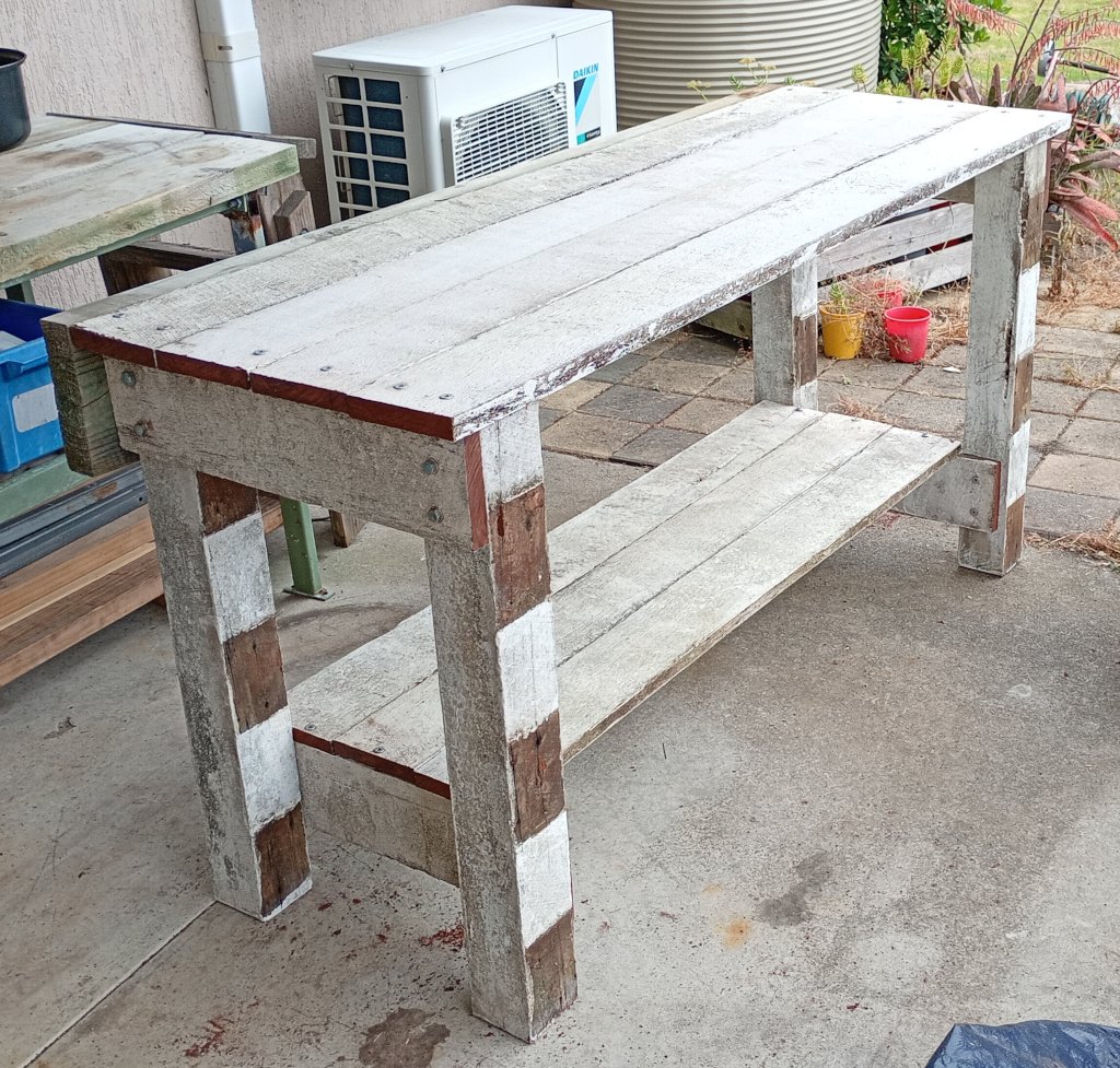A wooden bench stands on a concrete pad in the back yard of a house. The bench is mostly old white fence railings and fence posts with some freshly cut ends. It is pretty awesome.