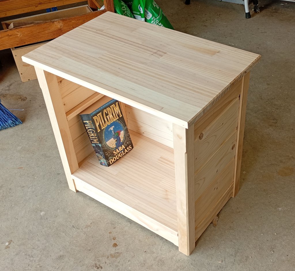 Photograph of a small bookshelf-cum-table. It is made of pine and spruce. Imagine a small occasional table but with pine paneling sides and back and a shelf about 70cm from the floor. A copy of Pilgrim by Sara Douglass is sitting inside. The bookshelf is sitting in the garage waiting to be finished.