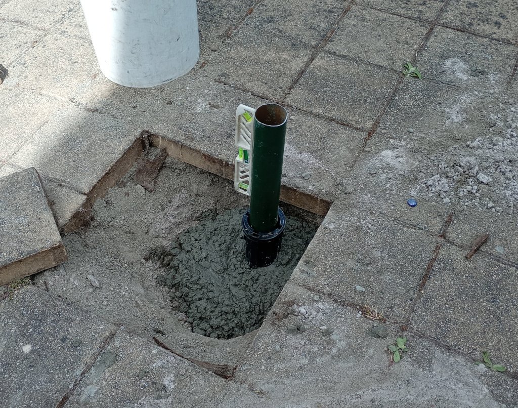 The picture shows the installation of a new socket for a clothes line. It is being concreted into the hole where the old clothesline was. A spirit level is attached to a length of the old post so that the clothesline will be vertical. 