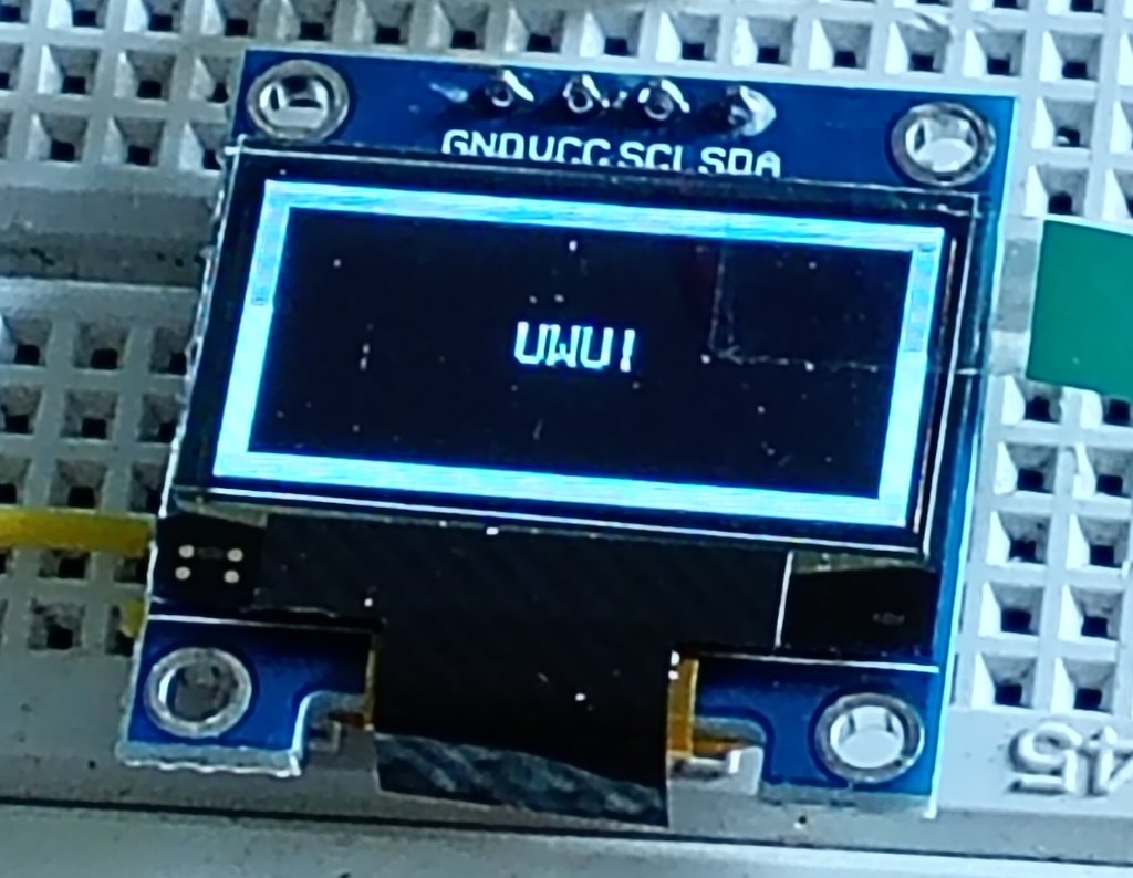 A picture of a mini OLED display plugged into a prototyping breadboard. It is square, though the actual display is smaller and rectangular. It has 4 connections at the top labeled gnd, vcc, scl and sda. The display shows a light border surrounding a black space with the word UWU! in the middle of the screen.