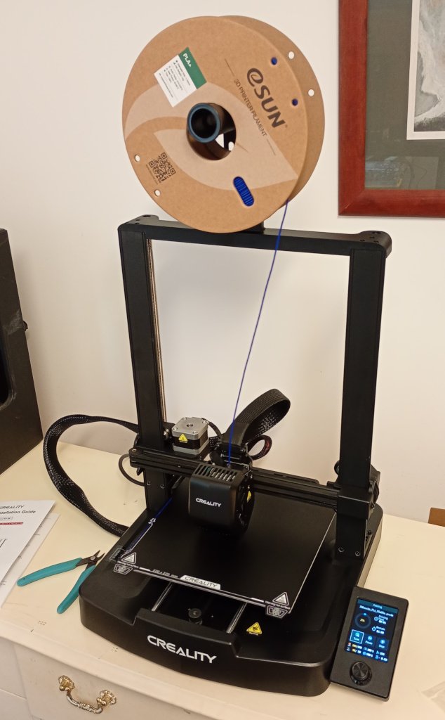 A 3D printer sitting on a chest of drawers. It is a Creality Ender 3 V3 SE with a gantry design. It is mostly black with the control panel on the front right corner of the printer. The printer is loaded with a spool of blue filament.