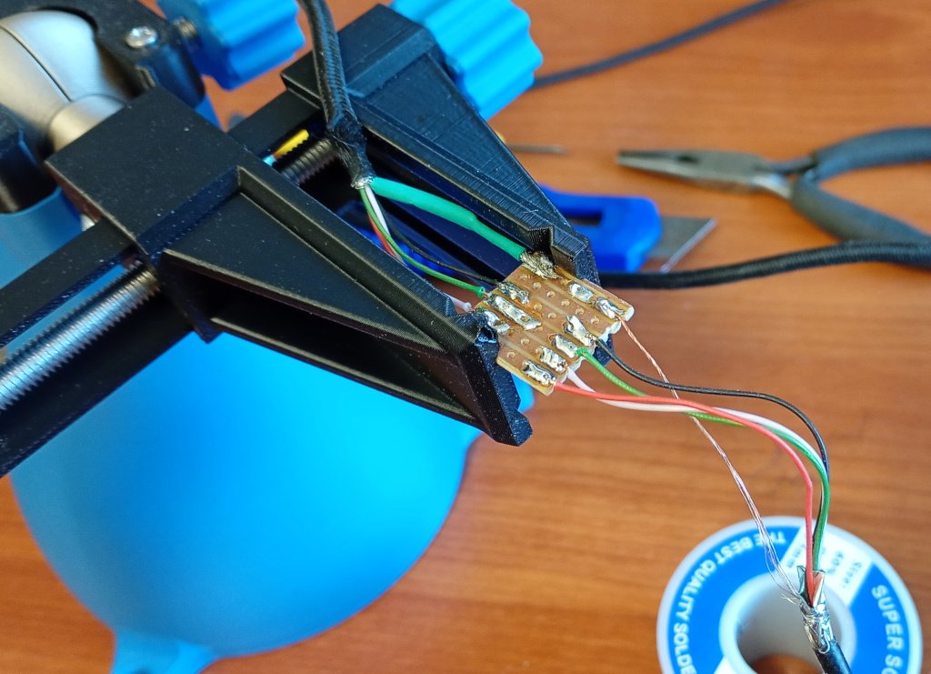 A picture of a tiny piece of prototyping board held in a PCB clamp. It has wires soldered to it, joining 2 sections of USB cable.