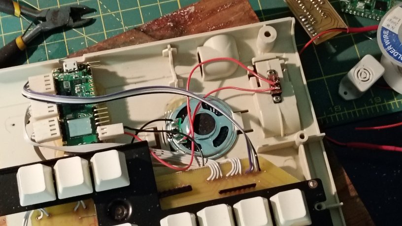 Picture of the inside of a keypad on a desk with random electronic junk. There are wires leading from a PCB to a microcontroller and a speaker and a switch.