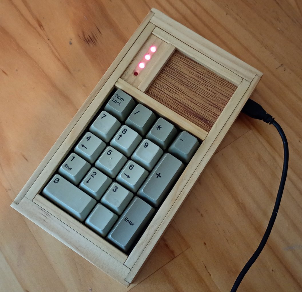 Close up view of a keypad. It is made of pieces of timber. The keys comprise a standard numpad. Above the numpad keys is a space divided vertically by a strip of wood. The left quarter of this space is occupied by four LEDs. On the right, the remaining space is filled by a piece of plywood. A USB cable is plugged into the keypad on the top-right side. 