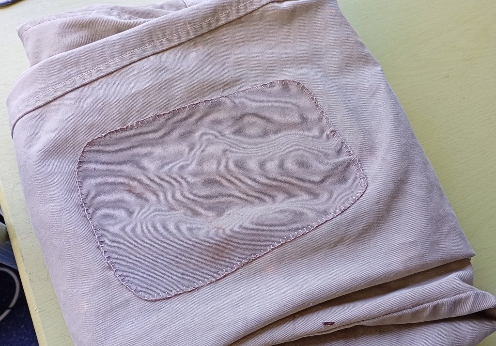 A picture of a pair of beige work pants, folded up on the table. The middle of the picture is dominated by a large patch on the knee, in the same colour as the pants. It is sewn onto the pants with a blanket stitch around the perimeter in matching cotton thread. I hope it works.