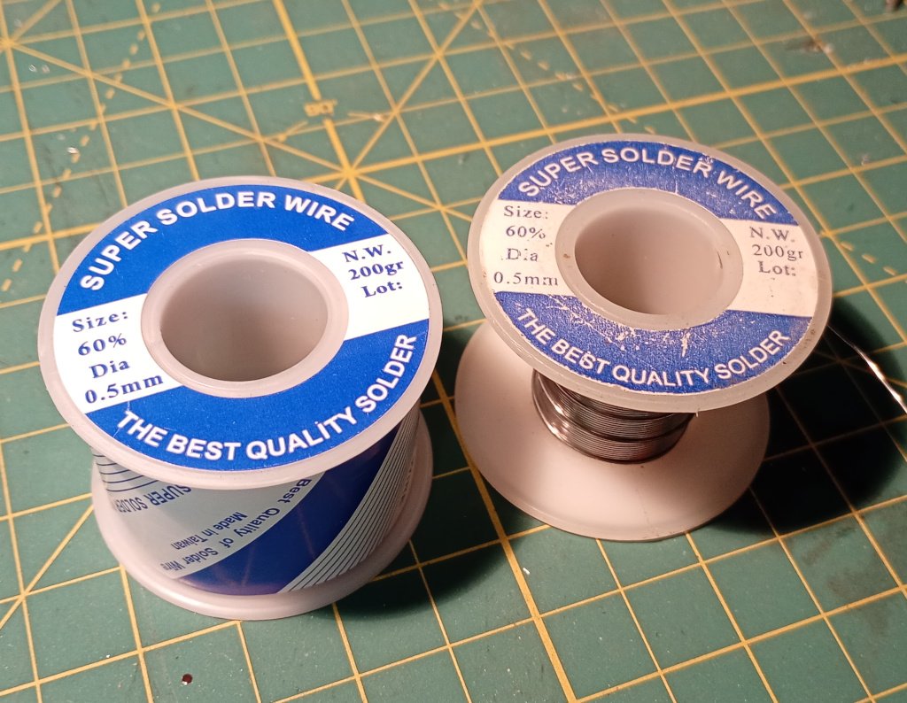 Two rolls of 0.5mm solder sitting on a craft mat. The one on the left is brand new. The one on the right is 20 years older and just about used up. The labels read 