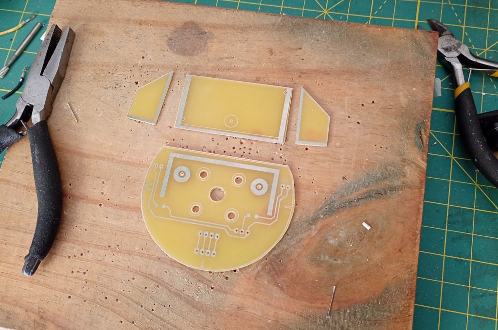 The picture shows a set of circuit boards laid out on a piece of wood. The semicircular circuit board contains traces for electronic circuits while the remaining boards are mostly bare. They will be soldered together to make a bracket for a sonar ranging module. The entire assembly will be mounted on a servo motor and the circular base has mounting holes to attach a servo horn.