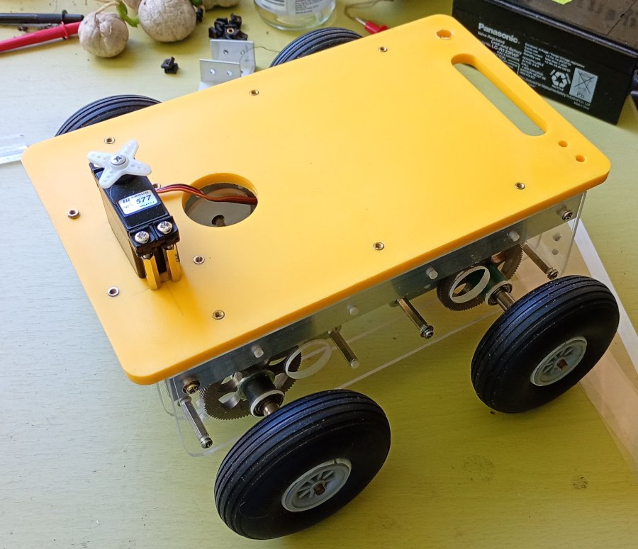 The picture shows a small wheeled robot base. It has a yellow plastic deck with a servo motor mounted on top. The sides are made from aluminium and acrylic. Gears can be seen connected to the axles that the wheels are attached to. The wheels are model aircraft wheels.