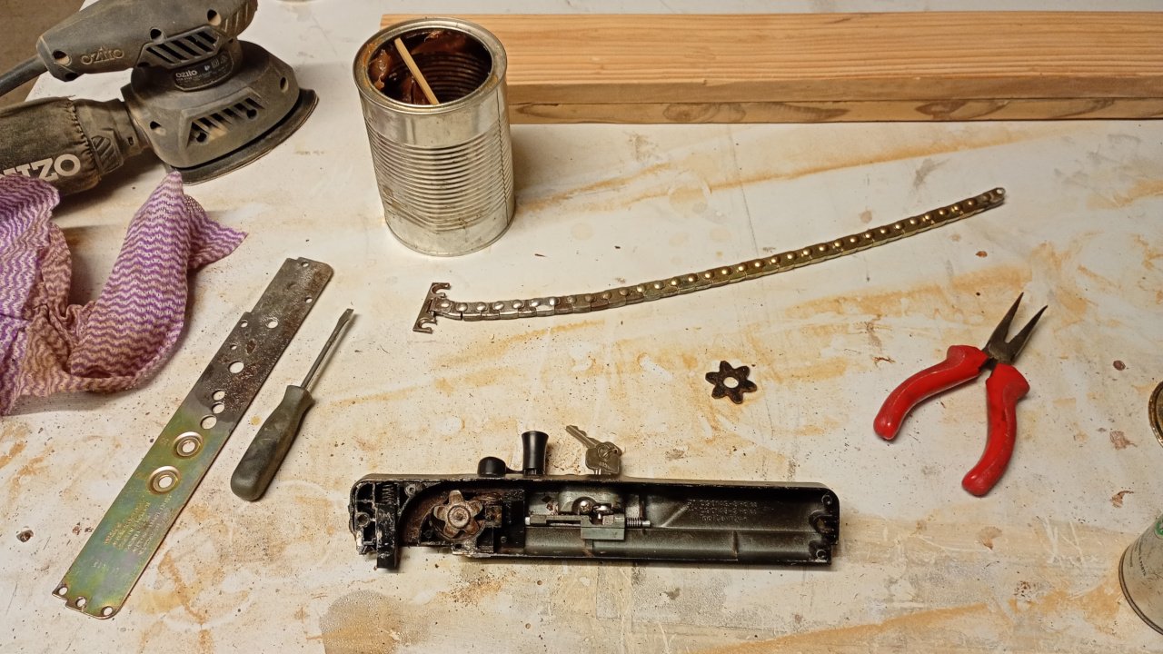 A dirty table with a number of things on it, principally a half disassembled Wilco window winder mechanism. The base plate has been separated from the body of the mechanism and is half covered in rust. The chain has also been removed and cleaned up . A tin of grease is at the top of the image and there's also a set op pliers and a sander (that I didn't use for this job. The plastic keeper cog is also visible.)