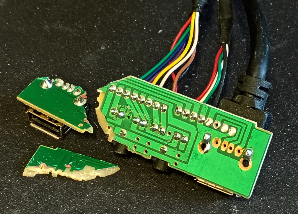 Close up picture of a broken PCB on a dirty mat. The PCB shows the copper side. There are many cables soldered to it. It is broken into three separate pieces and probably should be thrown away decently instead of being repaired. 