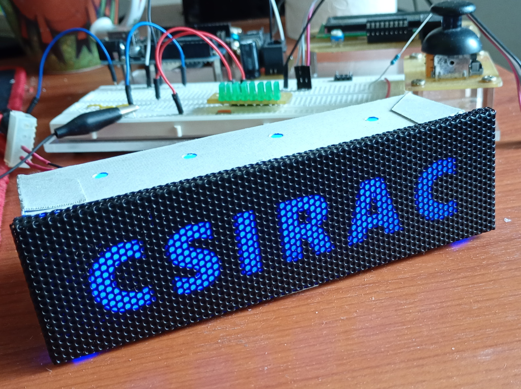 Close up of an illuminated computer name badge sitting on a wooden desk. It is sized to fit into a 5.25 inch drive bay. It is made of a black metallic mesh. The badge displays the name CSIRAC in bold Gill Sans blue illuminated letters. In the background is an electronics breadboard with wires, LEDs and some other electronic doodads.