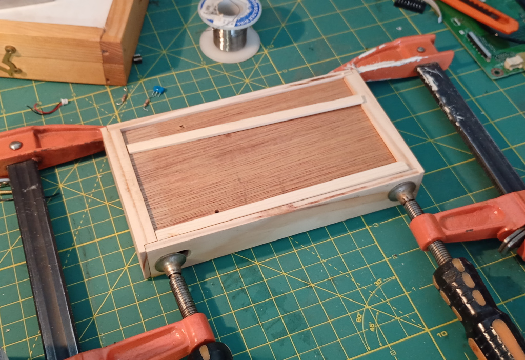 Picture of a wooden box being made on a hobby mat surrounded by electronic junk. The box is upside down with a bottom made of plywood. It is being clamped together at each end with orange clamps.