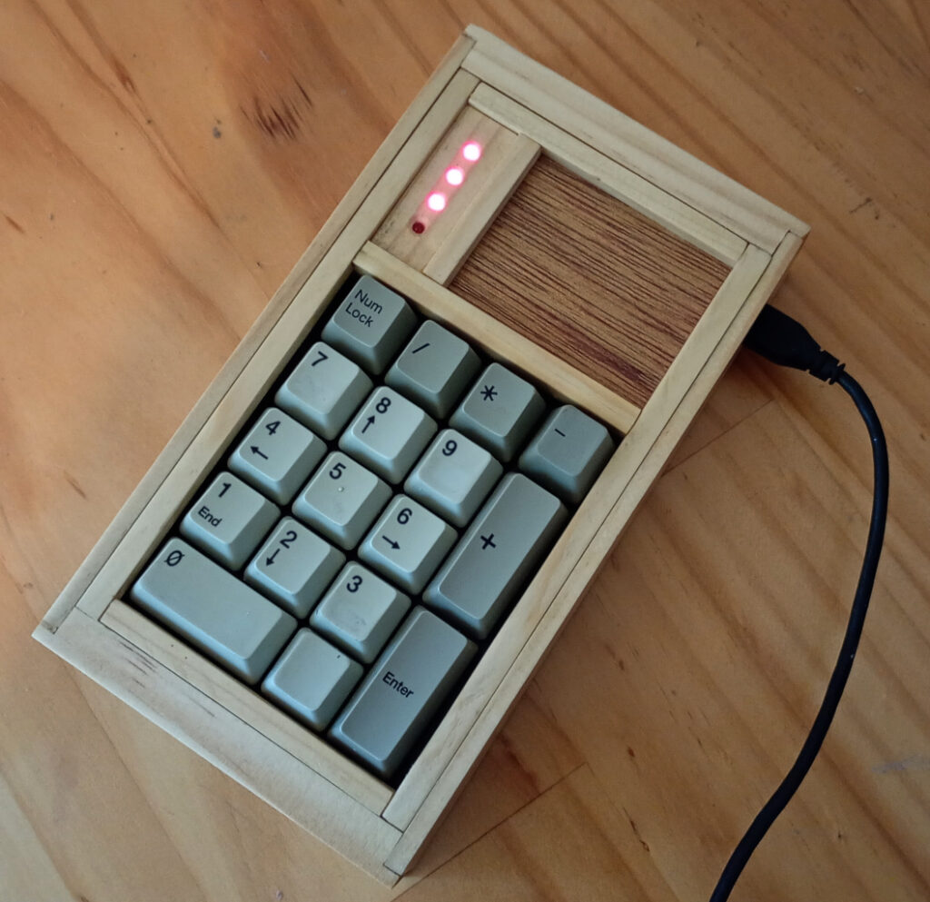 Close up view of a keypad. It is made of pieces of timber. The keys comprise a standard numpad. Above the numpad keys is a space divided vertically by a strip of wood. The left quarter of this space is occupied by four LEDs. On the right, the remaining space is filled by a piece of plywood. A USB cable is plugged into the keypad on the top-right side.
