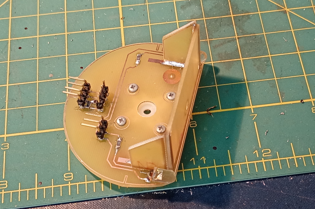 The finished servo deck, sitting on a craft mat. There are numerous components and connectors soldered to the semicircular base including SMD resistors and header pins and LEDs. A servo horn is screwed underneath the base. The mounting bracket for the sonar module is soldered in place creating a surface at 90° to the base to which the sonar module will be attached.