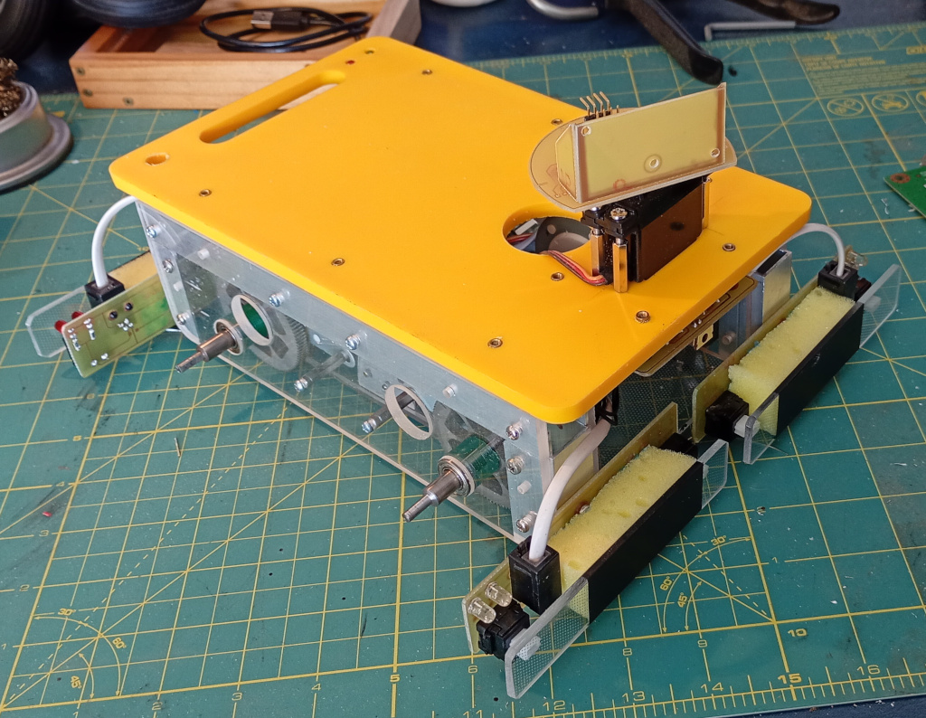 A mostly finished robot sits on a craft mat. It does not have wheels, but the sets of bumpers have been fitted to the front and rear. On the top deck a sonar turret made from circuit board material is mounted to a servo motor. The bumper modules are electrically connected to the control board with modular connectors.