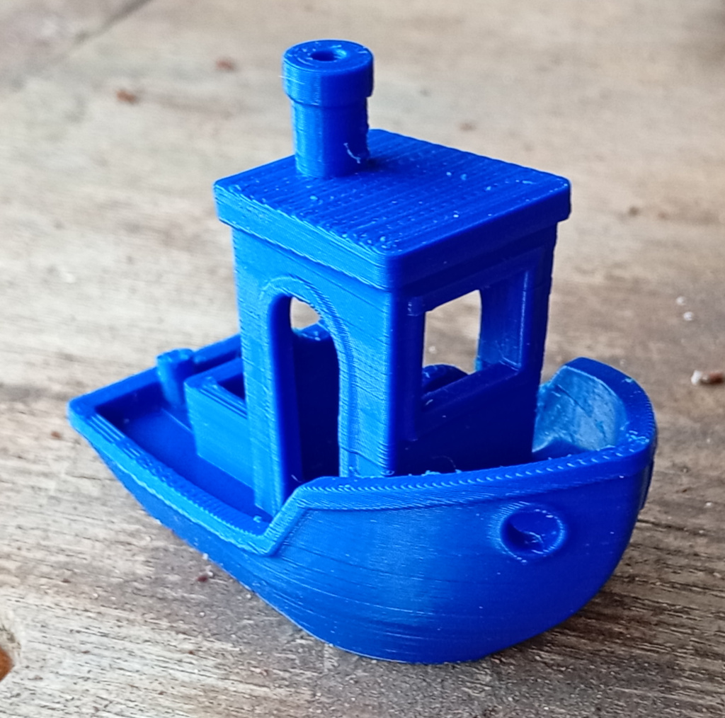 Close up of a benchy, a little boat model printed on the new printer. There are a few minor layer shifts visible and the remnants of some stringing but the overall print is sound. It is sitting on a wooden bench.