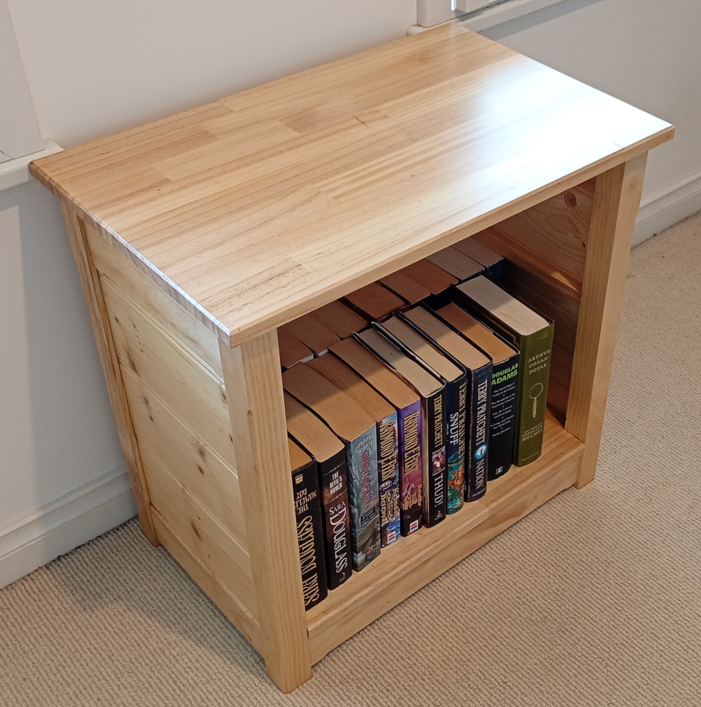 My bookshelf in the bedroom. It sits between 2 windows on pale beige carpet. It is full of large books by Douglass, Pratchett, Adams, Conan Doyle, Feist, Wurtz, and Jordan.