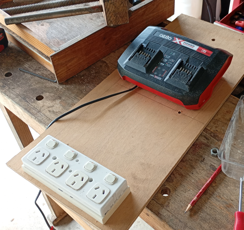 A length of plywood sits on a wooden bench. At the top is an Ozito battery charger and at the bottom is a 4-way power outlet that the charger will plug into.