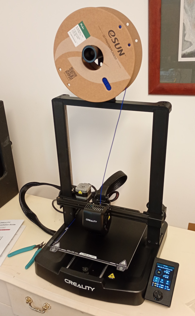 A 3D printer sitting on a chest of drawers. It is a Creality Ender 3 V3 SE with a gantry design. It is mostly black with the control panel on the  front right corner of the printer. The printer is loaded with a spool of blue filament.