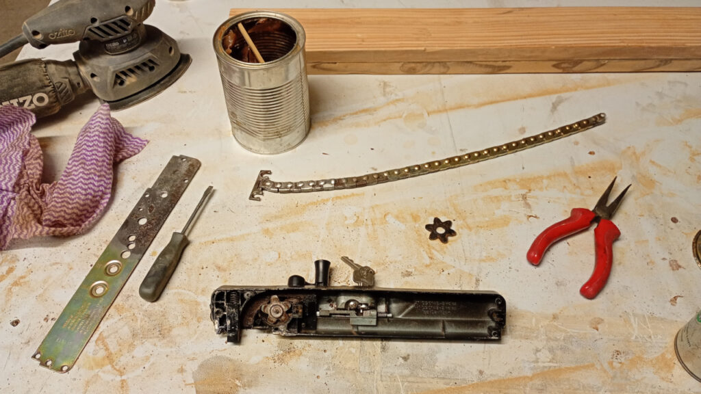 A dirty table with a number of things on it, principally a half disassembled Wilco window winder mechanism. The base plate has been separated from the body of the mechanism and is half covered in rust. The chain has also been removed and cleaned up . A tin of grease is at the top of the image and there's also a set op pliers and a sander (that I didn't use for this job. The plastic keeper cog is also visible.)