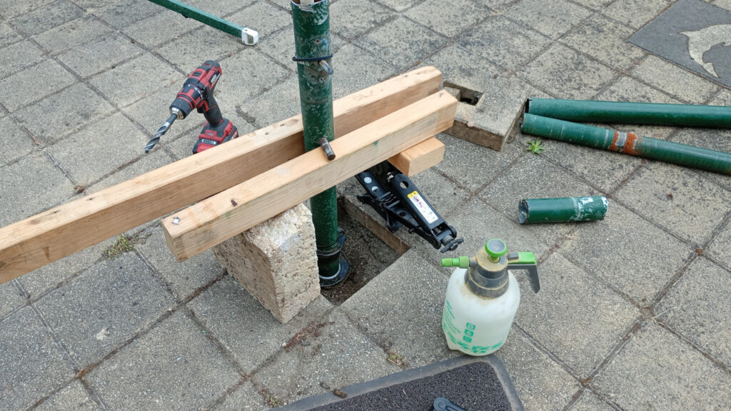 A backyard paved area. In the middle is the remnants of a rotary clothes line. It is surrounded by some tools. A metal pin has been inserted through the middle of it and lengths of pine wedged underneath, sitting on a limestone block at one end and a car jack at the other. Some other bits of the clothes line are also sitting on the ground.