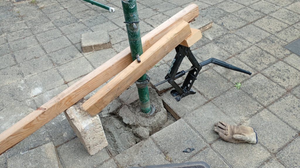 The jack has been lifted up and the wooden beams are at an angle. The clothesline has been lifted out of the ground along with the concrete footing. There is also a rigger's glove.