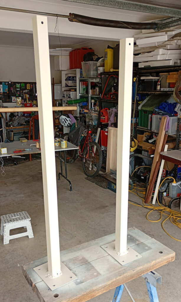 The picture shows the inside of a cluttered garage. In the foreground, two metal posts made of galvanised steel tube are standing on a sawbench after being spray painted. 