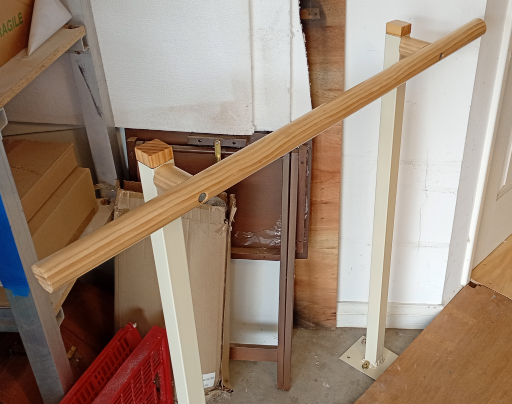 The picture shows a closeup of a wooden railing made from round timber. It is attached to steel posts bolted onto a concrete garage floor. There is a shelf to the left of the image and a pile of random garage stuff.