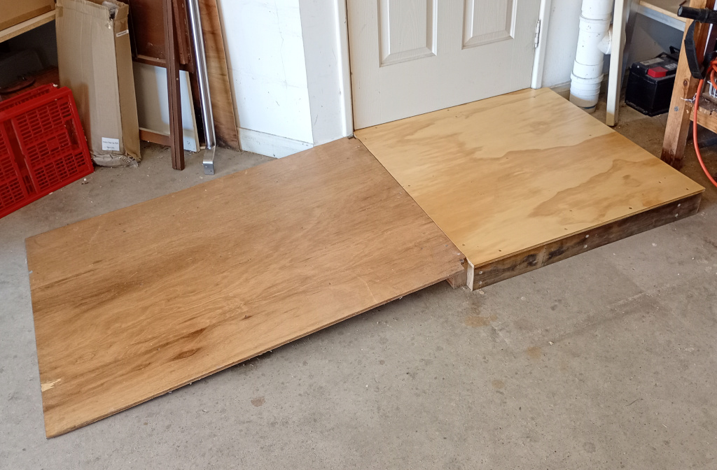 Picture shows part of a garage with a wooden accessibility ramp. The door to the rest of the house leads to a plywood square platform and a plywood ramp leads off to the left of the image to the rest of the garage. There are some other bits of garage stuff.