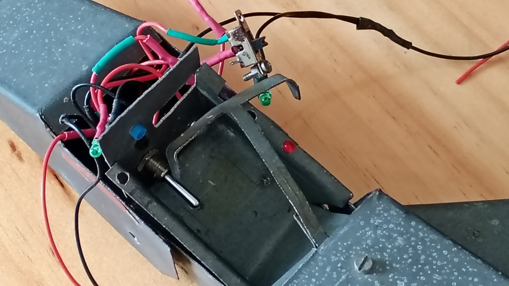 Closeup of the cockpit area of the toy jet before reassembly. A tangle of red and black wires and LEDs can be seen coming from behind the instrument panel as well as a slide switch. The main power toggle switch can also be seen. The metal is covered in white spots of corrosion.