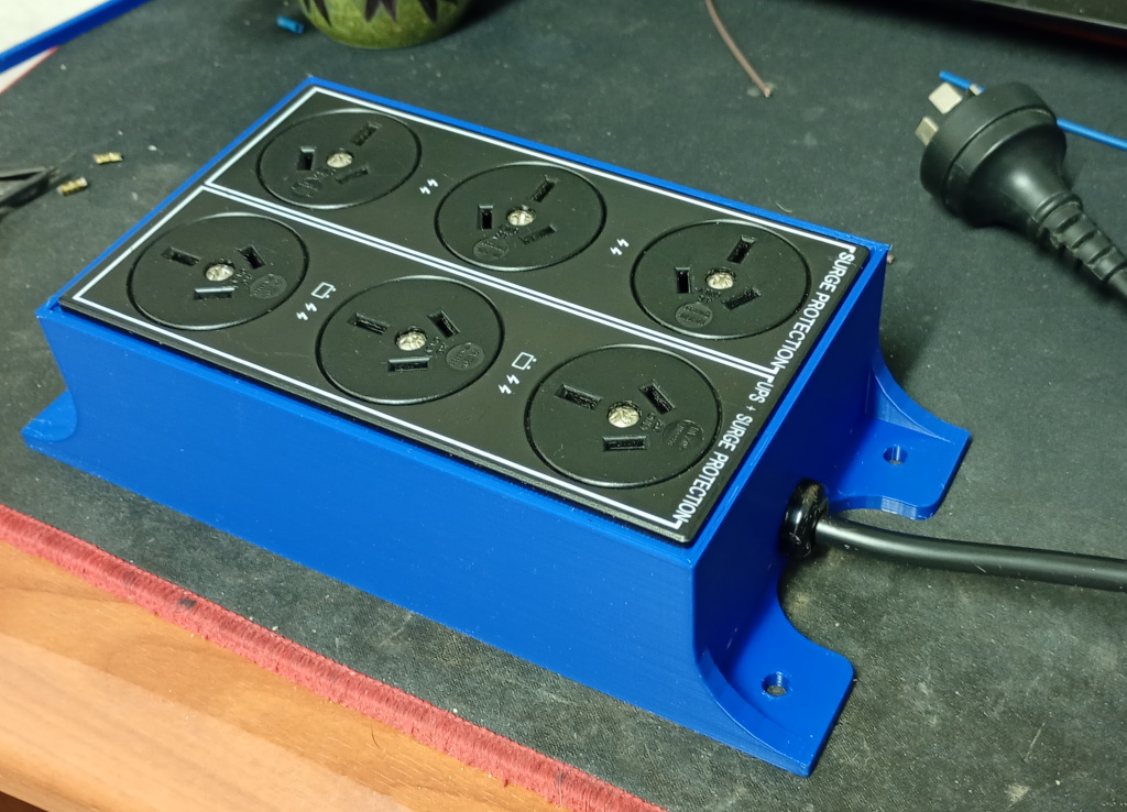 The finished power board is sitting on my desk mat. The panel of 6 mains plugs has been clipped into place in the enclosure and the black mains cord has been secured with a strain relief clamp in the provided hole. It looks pretty awesome.