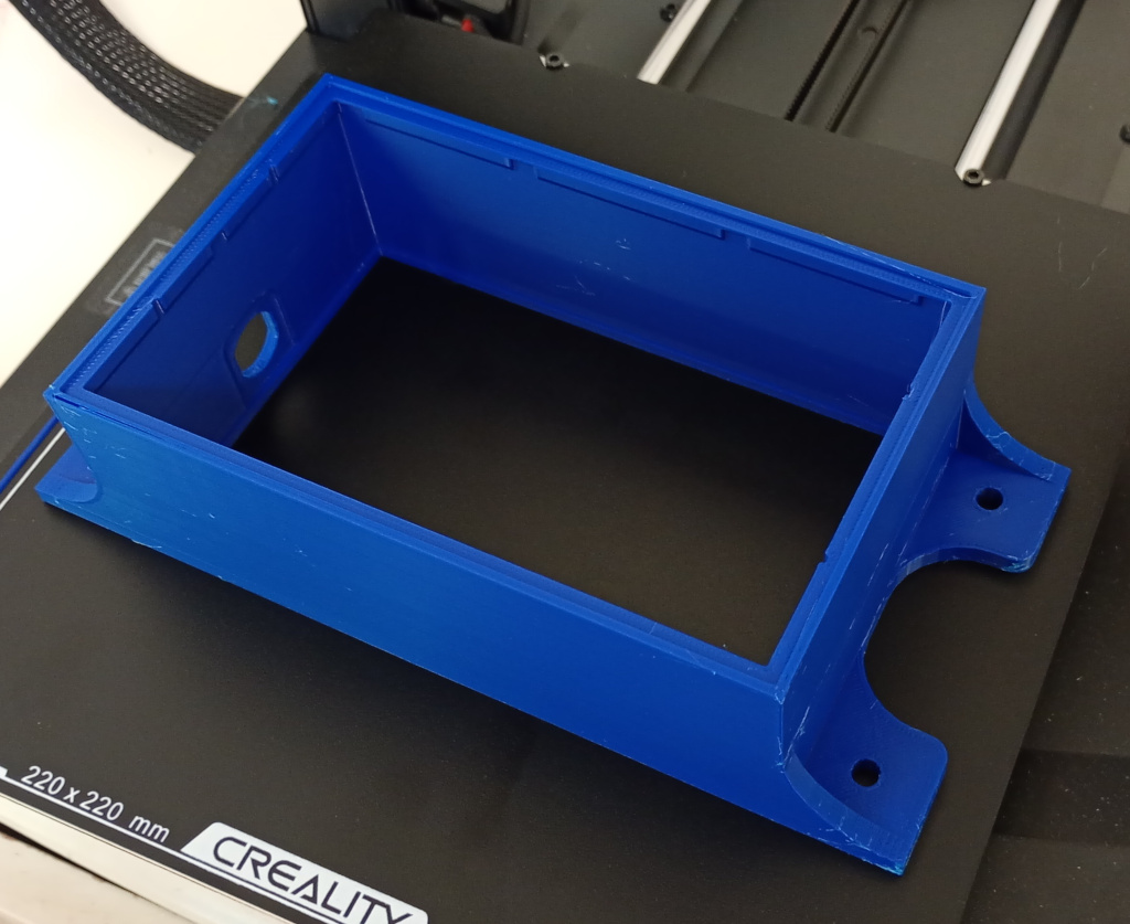 The 3D printed enclosure, sitting on the print bed of a 3D printer. The enclosure is printed in blue plastic, largely rectangular in shape but with mounting tabs on the short edges. The short edge on the left hand side includes an opening to allow for a cable clamp.   