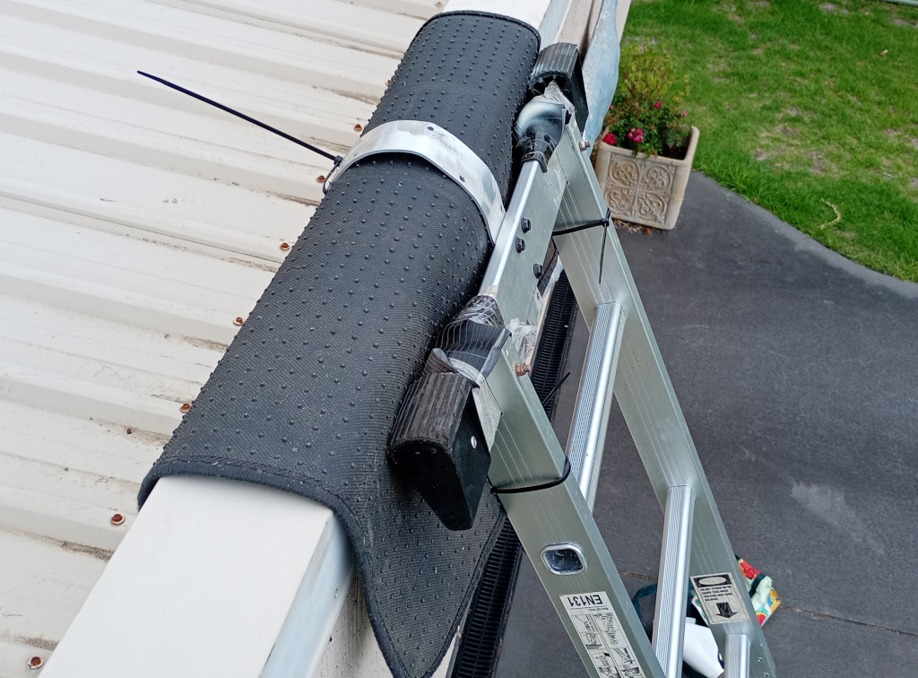 The ladder is leaning against the parapet of the low roof with the aluminium hook securing it in place. There is a car floor mat draped over the parapet, it's actually zip tied to the ladder, to protect the surface of the metal cladding of the house